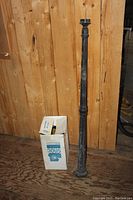 Full view of the black metal outdoor post light fixture standing vertically against wooden background with a branded box adjacent.