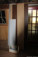Several flat shelf boards in white, brown, and grey leaning against a wall with a box of metal brackets on the floor.