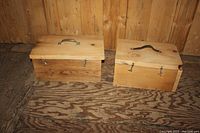 Two small wooden boxes made from natural wood with curved metal handles on top and hook latches on front, placed on wooden surface and against wood panel background.