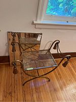 Metal bench with decorative tassel ends and mesh seat, bronze/antique gold finish, photo showing bench angled to camera with wooden floor and white wall background.