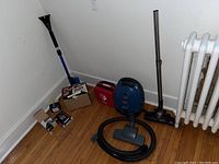 Full lot showing Bissell vacuum with hose and floor nozzle, red Philips Heartstart defibrillator, small red Shark hand vac, black lock box and some boxed deadbolts on hardwood floor by radiator against white wall.