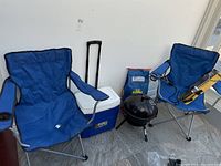 Two blue collapsible camp chairs, an Igloo wheeled cooler, portable black kettle BBQ grill, and bag of charcoal briquettes arranged on tiled floor against white wall.
