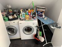Photo of laundry area with front-loading washer and dryer, various cleaning sprays and supplies on top, ironing board with iron and steamer box, and mop with bucket beside them.