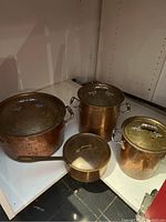 All four copper cookware pieces grouped inside a cabinet showing exteriors and lids.
