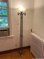 Full view of the floor lamp standing in a room, showing metal body and frosted glass shade lit with light on.