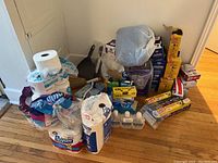 Wide view showing most household supply items stacked on floor including tissue paper, bags, wraps, hand sanitizer, and dust pans.