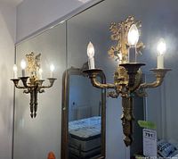 Photo showing one gold-colored metal light sconce with three candle-style bulbs mounted on a wall near a mirror causing reflection.