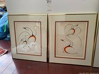Pair of framed signed prints showing abstract designs with black line work and gradients of orange to yellow against cream backgrounds, in gold frames with double matting.