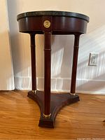 Side view of round side table showing three slender wooden legs with decorative gold-tone accents and triangular wooden base, wood stained dark cherry or mahogany.