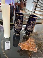 Photo showing two blue tinted glass vases with gold detailing and etched Asian village scenes, a translucent abstract ashtray and white pillar candle on glass table.