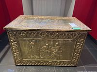 Front and side angled view showing embossed brass panel with classical figures and floral border.