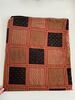 Folded fabric with a grid pattern in rust, brown, and black squares, showing textured prints with hearts, zigzags, and other motifs.