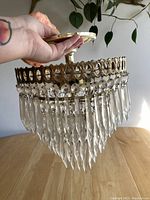 Side view held by hand showing brass ring frame with open oval cutouts and hanging transparent faceted crystal prisms forming a tapered shape.