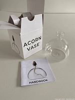 Photo showing the clear acorn shaped glass vase outside of its white box with Acorn Vase text and a handbook lying flat