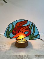 Front view of the handmade goldfish stained glass fan-shaped table lamp illuminated, showcasing the red goldfish and green plants on blue background, mounted on wooden base.