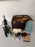 Full lot view showing microscope, wooden case with compartments for accessories, multiple glass bottles, cylindrical optical components, brush, clear glass tube, and instruction manual.