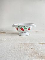 Front view of white enamel colander with bright red strawberries and green leaves design, showing side handles and pedestal base.