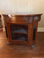 Angled front view showing circular mahogany top, brass lion head drawer pulls, wire-mesh cabinet doors, and brass claw feet
