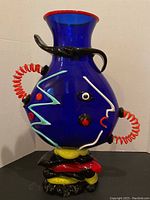 Front view of cobalt blue vase with red rim and black twisted handle, showing abstract face details.