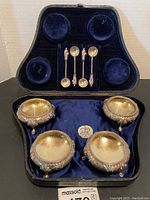 Open black case with blue velvet interior containing four ornate silver salt cellars on bottom and four silver salt spoons on top
