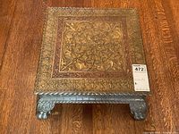 Top view showing square brass stool with ornate raised floral and scrolling pattern and claw feet visible beneath