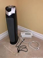 Side front view of Germguardian air cleaner tower with power cords and a small white heater vent nearby on floor.