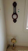 Photo showing the wall-mounted wooden barometer and the metal lamp positioned below it on the floor near a wall outlet.