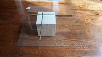 Top-down view of square glass top resting on a stone cube base with brass crossbars on hardwood floor.