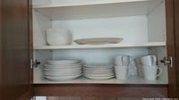 Photo of various stacked white dinnerware including plates, bowls, and mugs in a cabinet showing quantity and condition.
