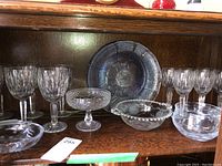 Shelf showing stemmed crystal wine glasses, small pedestal bowls, round plate with cut designs in wood cabinet