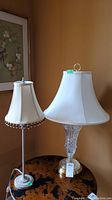 Two lamps side by side on a dark round table against a mustard-colored wall. Smaller lamp has a fabric shade with beaded trim and metal base, larger lamp has a plain white shade and glass/brass base.