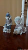 Two Lladro porcelain figurines on wooden floor, showing one standing child with bear cub and one seated child with bear cub.