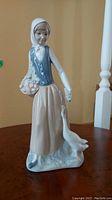 Front angle showing girl figurine with headscarf, basket of flowers, and goose at her side on base.