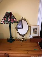 Photo of oval ornate mirror with brass-tone frame, floral lampshade lamp, framed print, and wooden tissue box holder on wooden surface against yellow wall.