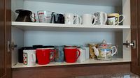 Two shelves with multiple mugs, a clear Pyrex measuring cup, and a floral decorative teapot with colorful lid handle.