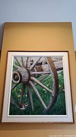 Framed signed painting showing a close-up of a wooden wagon wheel with chicken in green grass, hung on wall.