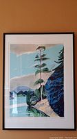 Framed art print mounted on a beige wall, showing a landscape with water, greenery, and a tall tree.
