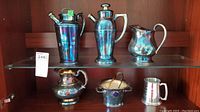 Five silver plate items displayed on glass shelf showing tarnished condition including cocktail shaker, coffee pot, pitchers, sugar bowl with tongs, and mug.