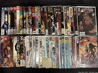 Photo showing 50 assorted comic books arranged in two rows on a white surface, many in protective plastic sleeves.