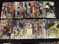 Photo showing 50 comic books laid out in two rows, featuring DC and Marvel titles including Superman, Green Lantern, Batman, and other mixed titles. Comics are in plastic sleeves with backing boards for protection.