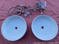 Two white round porcelain sinks with central drain holes displayed side by side with three metal faucets above them on a red textured fabric background.