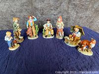 Group photo of 7 Homco Interiors figurines arranged on a dark cloth background showing various Western-themed characters in colorful painted ceramics including a seated man playing guitar, cowboys, woman in pioneer attire, and cattle figurines.