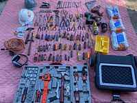 Photo showing a large assortment of hand tools including pliers, screwdrivers, wrenches, hammers, levels, helmets, tape measure, and an assortment of power tools in cases on pink quilted surface.