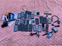 Wide shot showing a variety of cell phones laid out on a pink textured surface, including smartphones, flip phones, and older keypad models plus some accessories.