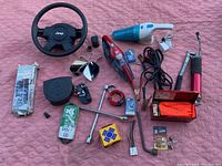 Top view of lot items including Jeep steering wheel, handheld vacuums, automotive light bulbs, and tools on pink background