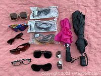 Overview of eleven pairs of sunglasses with various frame and lens styles, two umbrellas: one with pink cover and one with black carrying case on pink fabric