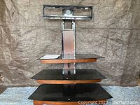 Full view of the TV stand showing three black tinted glass shelves, metal central mounting support, and wood-like trim.