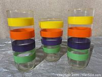 Three vintage glass cups with colored horizontal bands in yellow, orange, purple, and green arranged in a row on a gray surface