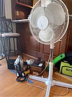 Photo showing the white pedestal fan along with shoeshine box, iron, flashlights, and part of the Nikon binoculars carrying case on the floor near stools and wooden cabinets.