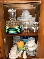 Full view of cabinet shelf with stacked Fiesta ware plates and cups in various colors, glass plates, white ceramic plates, glasses with floral patterns, and white ceramic salt and pepper shakers.